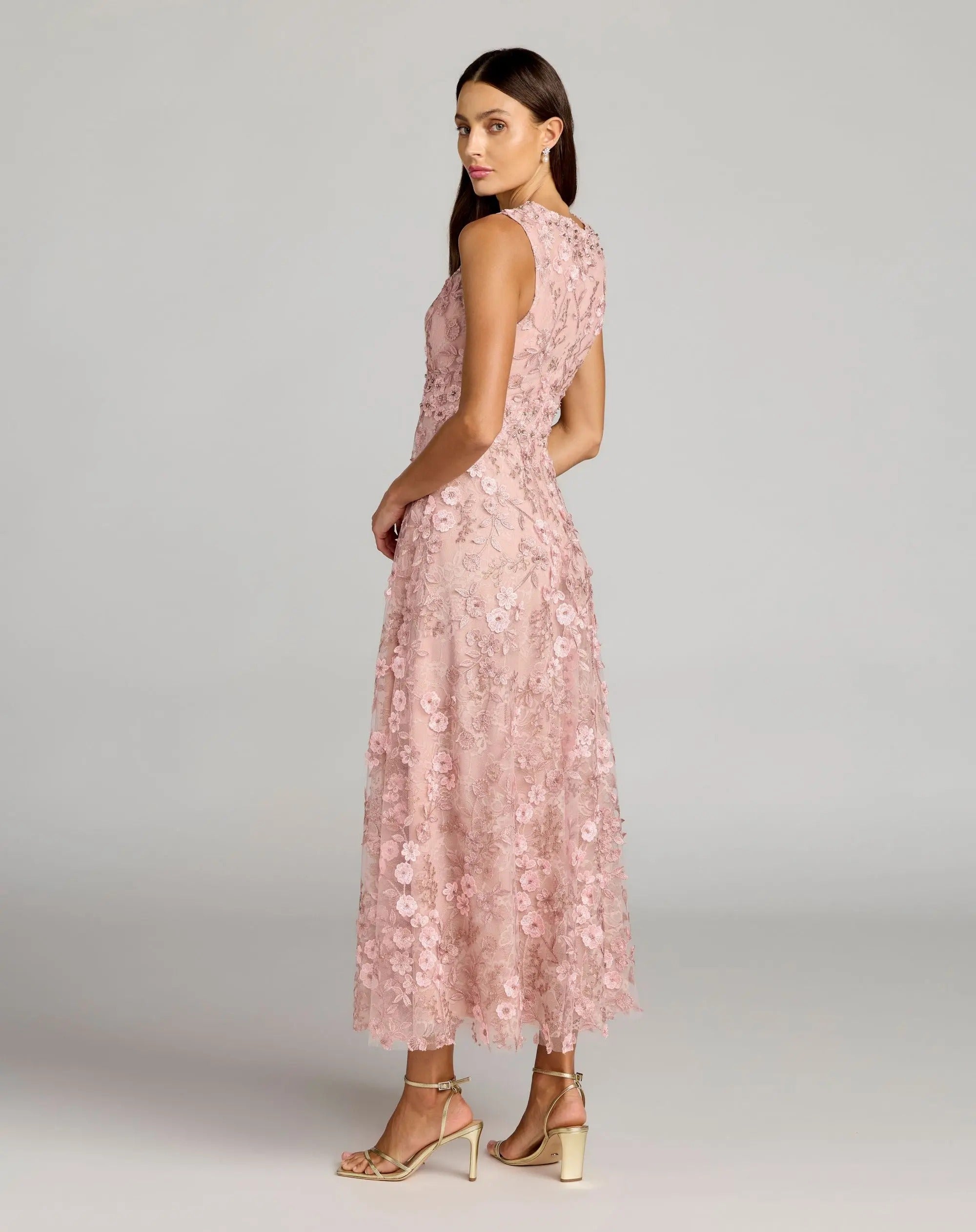 A woman stands sideways against a plain gray background, wearing the Mac Duggal 69192 tea-length party dress—a sleeveless light pink ensemble with a floral lace overlay, fitted bodice, and elegant A-line skirt—paired with ankle-strap heels.